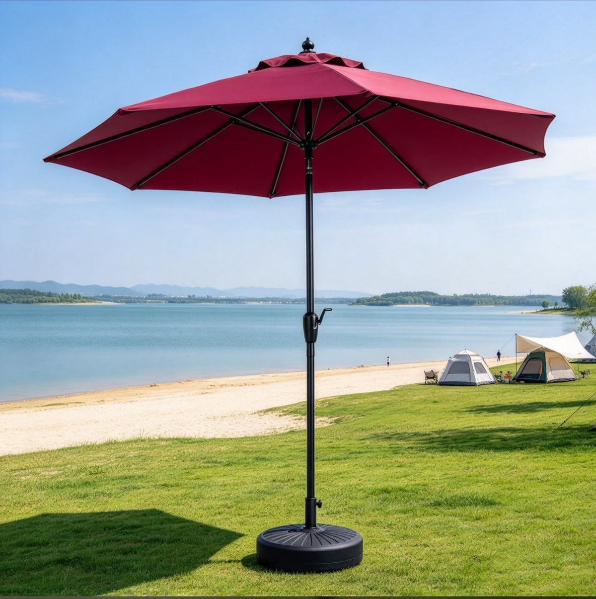 A large red patio umbrella with a black pole and weighted base stands on a green lawn by the beach, with water, sand, and tents in the background.