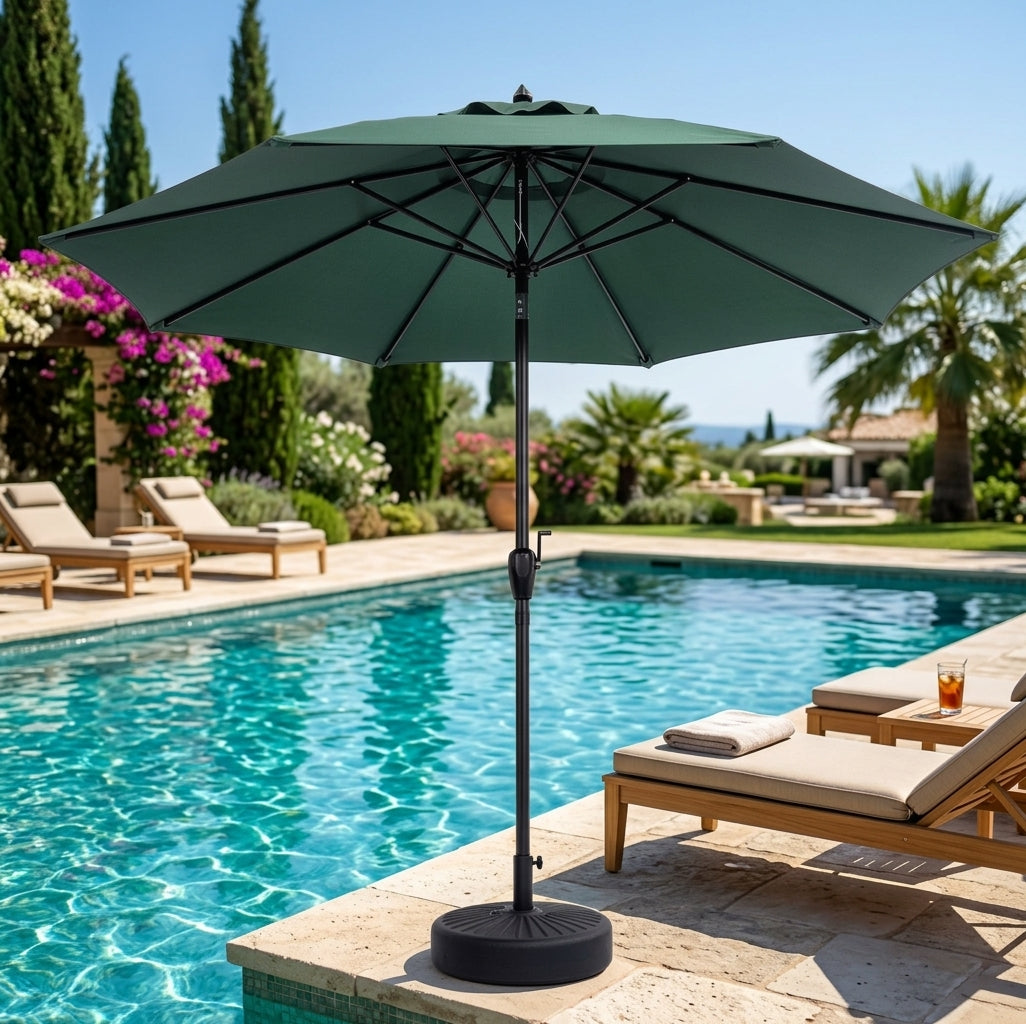 A large dark green patio umbrella with a black base and pole stands next to a swimming pool in a luxurious outdoor setting with lounge chairs, palm trees, and a clear blue sky.