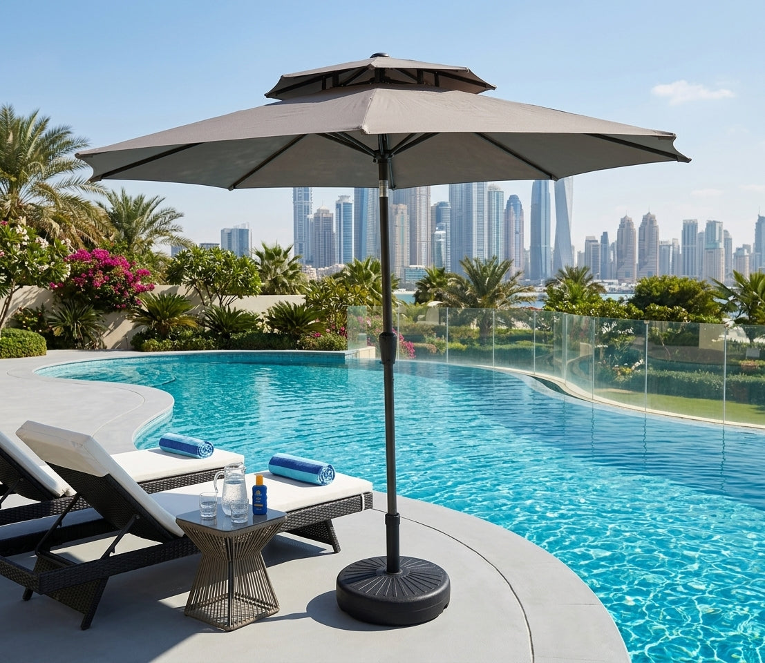 A large grey patio umbrella with a double-tiered canopy stands beside a swimming pool on a resort terrace, with lounge chairs, a small side table, palm trees, and a city skyline in the background.