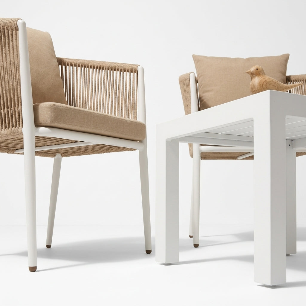A modern outdoor patio chair with a white frame and woven beige rope seat and back, shown next to a matching white table with a beige cushion and a small decorative bird figurine.